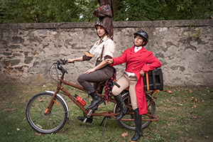 Two actresses, one in red waistcoat with tails and one with a theatrical horse head, pose on tandem bicycle on lawn in front of tall slate and stone wall.